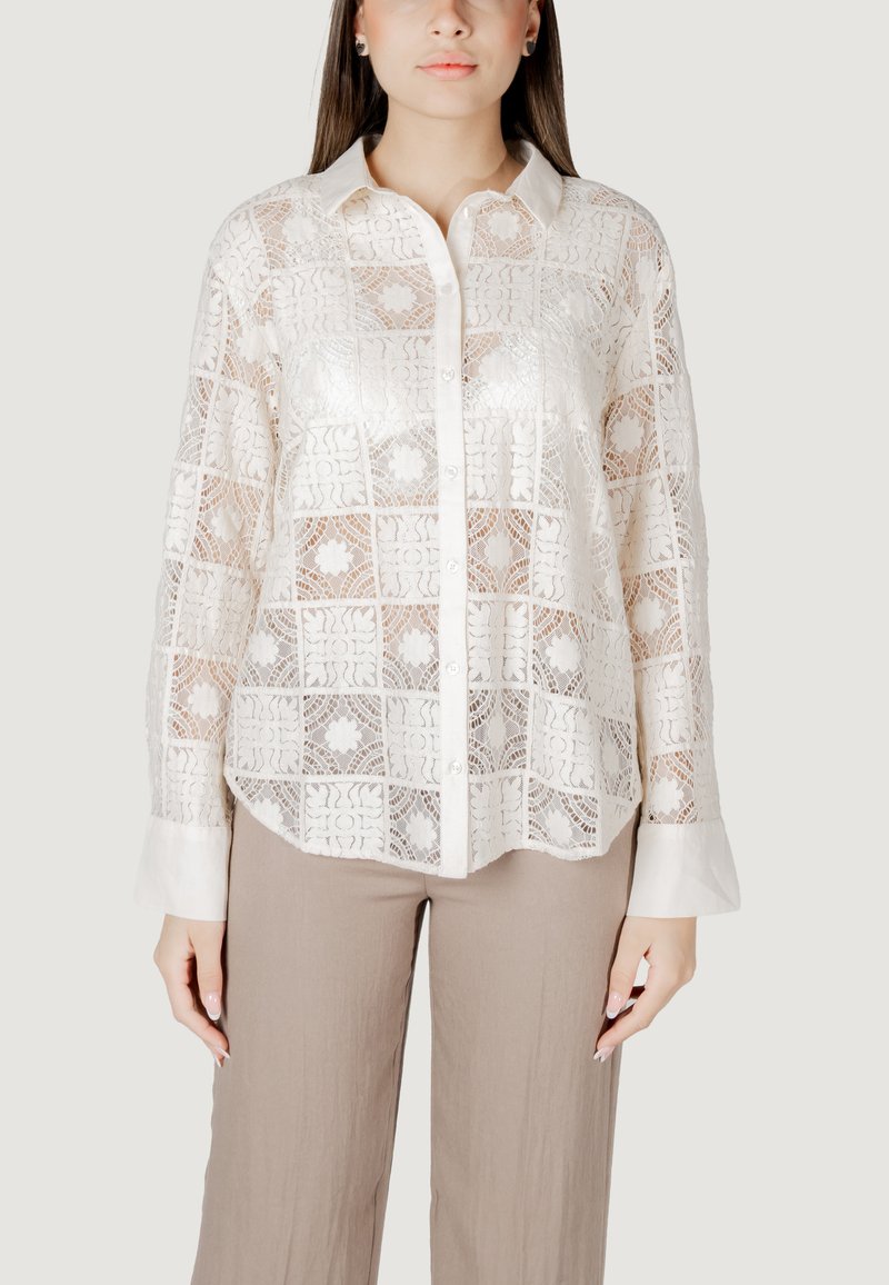 Valge pitsbluus keeruka ruutmustriga, millel on krae, nööpidega esiosa ja pikad varrukad. Kombineeritud beežide laiade pükstega.