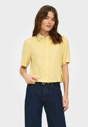 Ung kvinde med brunt hår iført en gul kortærmet skjorte med knapper, der ikke er stukket ind, over mørkeblå jeans, poserede mod en hvid baggrund.