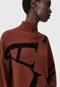 Brązowy sweter z golfem z czarnym wzorem logo, prążkowany kołnierz i mankiety, miękka tekstura, luźny fason oraz widoczne szwy na ramionach.