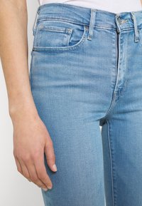 Vaqueros de mezclilla azul claro con un corte ajustado, diseño de cinco bolsillos y detalles de costura sutiles. Mano visible descansando sobre la tela.