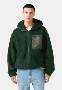 Zelená fleecová bunda s textúrovanou tkaninou, kapucňou, bočnými vreckami a zipsovým vreckom na hrudi so značkou. Nosená cez svetlomodré džínsy.