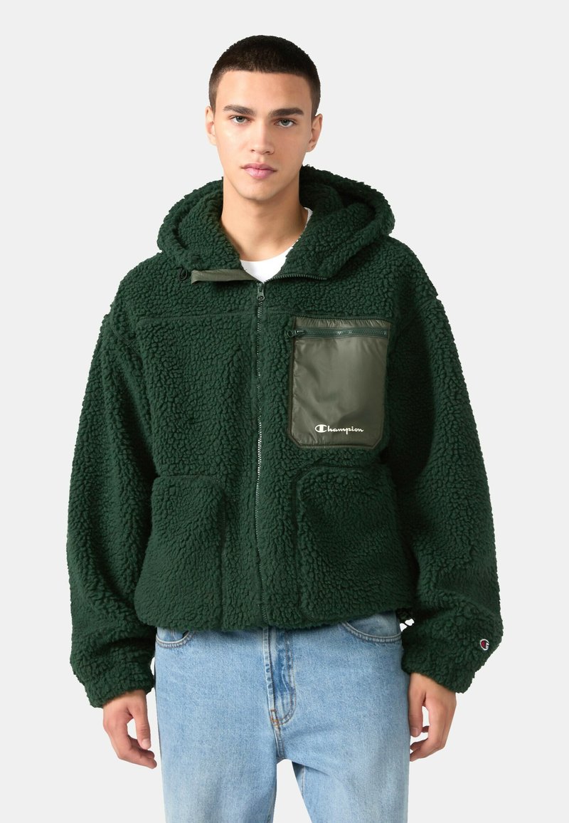 Zelená fleecová bunda s textúrovanou tkaninou, kapucňou, bočnými vreckami a zipsovým vreckom na hrudi so značkou. Nosená cez svetlomodré džínsy.