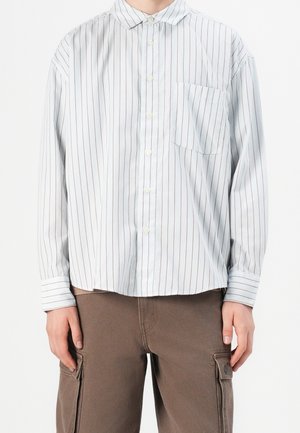 Person iført en hvid langærmet skjorte med tynde lodrette brune striber og brune cargobukser med forlommer.