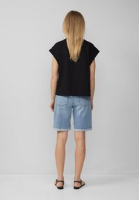 Schwarzes Kurzarmoberteil kombiniert mit hellblauen Jeansshorts mit umgeschlagenen Säumen, getragen mit schwarzen flachen Sandalen, von hinten gesehen.