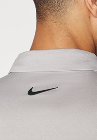 Pelēks polo krekls ar smalku tekstūru un baltu apkakli. Tam ir neliels melns Nike logs aizmugurē.