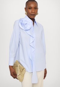 Ανοιχτό μπλε πουκάμισο με κουμπιά και μεγάλο διακοσμητικό λουλούδι από ύφασμα στον ώμο, με ρυτιτωμένο μπροστινό μέρος. Συνδυασμένο με μια χρυσή τσάντα ποιότητας.