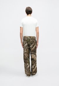 Στρατιωτικές παντελόνες με πατίνα καμουφλάζ σε καφέ και πράσινες αποχρώσεις, με άνετη εφαρμογή και ελαστική μέση, φορεμένες με ένα απλό λευκό πουκάμισο.