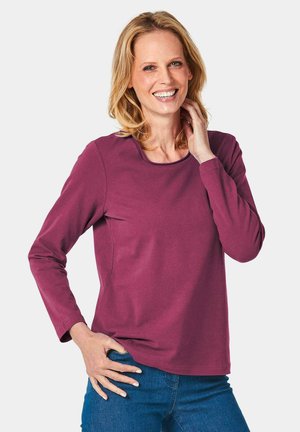 Langärmliges T-Shirt mit Rundhalsausschnitt aus gedämpftem burgunderfarbenem Stoff, das eine glatte Textur und dezente seitliche Nähte aufweist, kombiniert mit blauen Jeans.