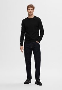 Sweater preto de manga longa feito de tecido suave, combinado com jeans de denim azul escuro e sapatos pretos com atacadores. Ajuste simples e moderno.