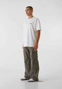 Biele bavlnené tričko s čiernym textom je doplnené olivovorozovými cargo nohavicami s viacerými vreckami a voľným strihom. Šedé tenisky dotvárajú celkový vzhľad.