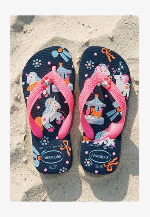 Tongs bleu marine pour enfants avec brides roses présentant des illustrations de licorne, carrousel, nœud et fleur sur sol sablonneux.