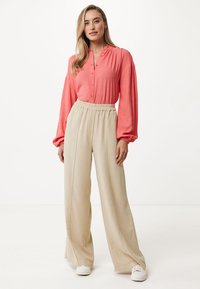 Korálová košile na knoflíky s dlouhými rukávy a elastickým pasem, kombinovaná s béžovými kalhotami s širokými nohavicemi, doplněná bílými teniskami na neutrálním pozadí.