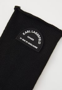 Svart strikket tilbehør med en rund, opphøyet logo-patch som viser "KARL LAGERFELD," "PARIS," og en adresse i hvite bokstaver.