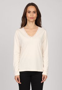 Haut crème à manches longues avec un décolleté en V, orné d'une délicate bordure en dentelle. Tissu doux au toucher lisse. Associé à un pantalon noir.