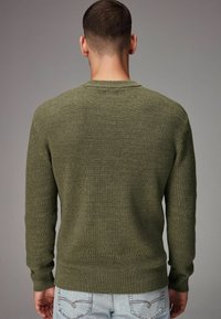 Olijfgroen gebreid trui met een ronde hals, geribbelde textuur en lange mouwen, gedragen met lichtblauwe jeans. Geen zichtbare patronen of versieringen.