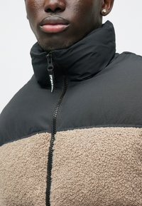 Jachetă din fleece neagră și bej cu guler înalt, secțiune inferioară texturată și fermoar frontal. Are panouri din țesătură contrastante și etichetă logo.