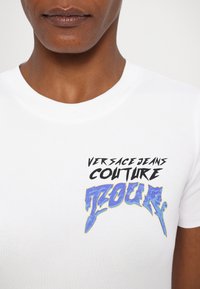 Balta medvilninė marškinėliai su grafiniu spaudiniu, kuriame užrašas "VERSACE JEANS COUTURE" juoda spalva ir "TOUR" mėlyna spalva su geltonais akcentais.