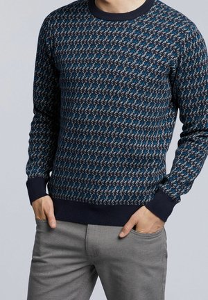Mężczyzna ubrany w sweter w granatowo-niebiesko-szary wzór z granatowymi mankietami, do tego szare spodnie, ręce w kieszeniach, na jednolitym tle.