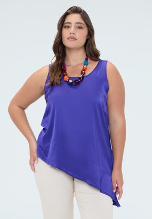 Vrouw met golvend haar, gekleed in een paarse mouwloze asymmetrische top, beige broek en een grote kleurrijke kralenketting, staand met één hand op de heup.
