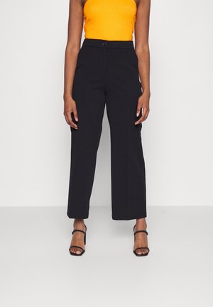 Femme portant un pantalon noir taille haute à jambes larges, un haut orange sans manches et des talons ouverts noirs, debout devant un fond uni.