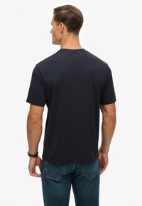Mann trägt ein schlichtes schwarzes T-Shirt und blaue Jeans, steht mit dem Rücken zur Kamera vor einem weißen Hintergrund.