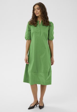 Femme aux cheveux bruns bouclés portant une robe verte mi-longue à manches bouffantes et des escarpins noirs à talons bas avec bride arrière, debout sur un fond blanc.