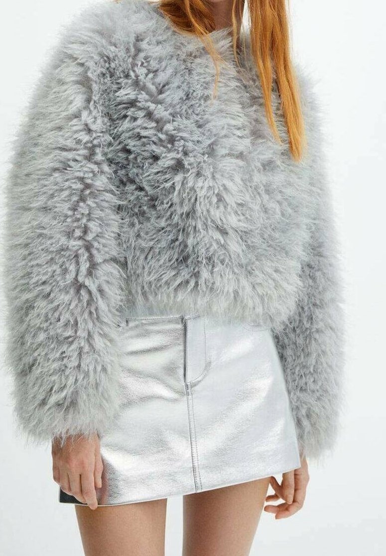 Femme portant un pull gris clair duveteux et une mini-jupe argentée brillante, debout devant un fond uni.