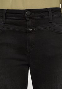 Jeans de denim negro con un diseño ajustado, estilo clásico de cinco bolsillos y detalles de costura sutiles, con una etiqueta de marca en la cintura.