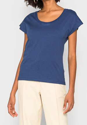 Frau trägt ein marineblaues, kurzärmliges T-Shirt mit U-Ausschnitt und hellbeige Hose, steht vor einem schlichten Hintergrund.