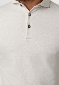 Vaaleanbeige pehmeästä kankaasta valmistettu poolopaita, jossa on kaulukset, kolme mustaa nappia ja hienovarainen tekstuuri.