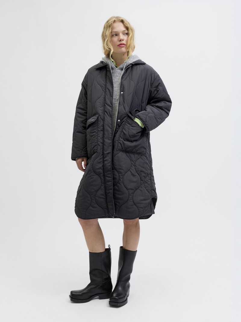 Gesteppten schwarzer knielanger Mantel mit Kapuze, großen Fronttaschen und einem Reißverschluss, über einem grauen Hoodie und schwarzen klobigen Stiefeln getragen.