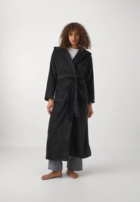 Črn kopalni plašč s kapuco iz mehke tkanine, z dolgimi rokavi, pasnim trakom in dvema sprednjima žepoma. Nošen preko sivega domačega oblačila.