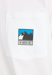 Bela majica s črno in modro kvadratno logotipom gora ter besedilom "THE NORTH FACE", ki je nameščeno na levi strani žepa.
