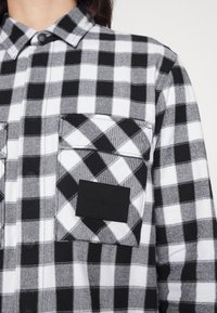 Koszula w czarno-białą kratę z kołnierzykiem na guziki, dwoma kieszeniami na piersi oraz czarną metką z logo. Materiał wydaje się mieć strukturę tkanej tkaniny.