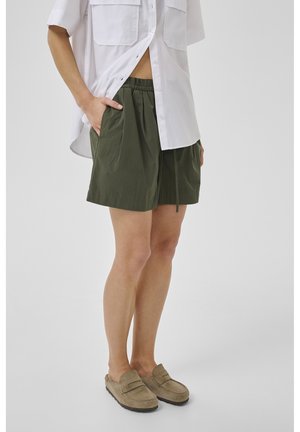 Personne portant un short ample vert olive avec une ceinture élastique et des chaussures beige à enfiler, associée à une chemise blanche à boutons, debout.