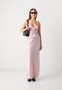 Abito di raso rosa con spalline sottili, dettaglio frontale attorcigliato e silhouette fluida. Abbinato a accessori neri e occhiali da sole.
