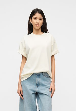 Frau mit langen dunklen Haaren, die silberne Creolen, ein locker sitzendes weißes T-Shirt mit hochgekrempelten Ärmeln und hellblaue hoch taillierte Jeans trägt, steht vor einem weißen Hintergrund.