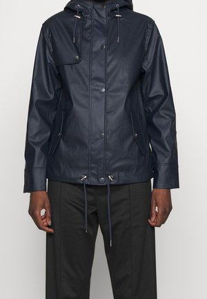 Tmavě námořnická dešťová bunda se zapínáním na druky, kapucí se stahovacími šňůrkami, klopovými kapsami a nastavitelným lemem, nošená přes černé kalhoty.