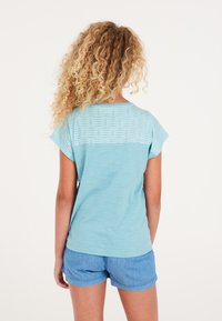 Camiseta azul claro con mangas cortas tipo capa y un respaldo con textura y patrón, combinada con pantalones cortos de mezclilla. El tejido parece suave y ligero.