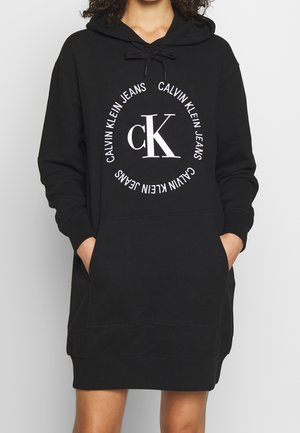 Czarna sukienka z kapturem Calvin Klein Jeans z białym okrągłym logo i rękami w przedniej kieszeni, noszona przez osobę widoczną od szyi do połowy uda.