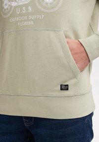 Sudadera verde claro con un bolsillo frontal, dobladillo acanalado y logo bordado. Tejido de mezcla de algodón suave con detalles de costura en contraste.