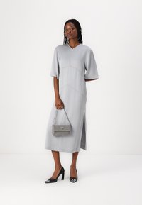 Hellgraues, kurzärmliges Midikleid mit V-Ausschnitt, seitlichen Schlitzen und geometrischen Nahtdetails. Kombiniert mit einer bejeweled grauen Handtasche und schwarzen Absätzen.