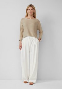 Beige gestrickter Pullover mit Dreiviertelärmeln, kombiniert mit weißen, weiten Hosen. Das Model trägt goldfarbene Ohrringe und beigefarbene Sandalen.