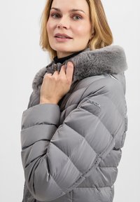 Giacca a sacco grigia con design trapuntato, dotata di un morbido colletto in pelliccia sintetica grigia e logo sul braccio sinistro, chiusura con zip e colletto alto.