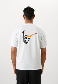 Bílý bavlněný tričko s velkým černým grafikou s zelenými a oranžovými akcenty na zádech. Krátké rukávy a volný střih.