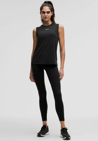 Czarny bezrękawnik sportowy z okrągłym dekoltem i logo, w połączeniu z dopasowanymi czarnymi legginsami i czarnymi sneakersami. Gładka tekstura.