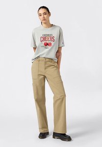 Camiseta de color gris claro con texto gráfico rojo e ilustraciones de cerezas, combinada con pantalones cargo beige con múltiples bolsillos y botas negras.