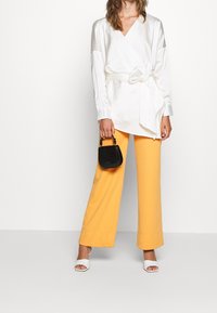 Blouse cache-cœur en satin blanc à manches longues, associée à un pantalon jaune à jambes larges. Accessoires comprenant un petit sac à main noir et des sandales blanches à talons.