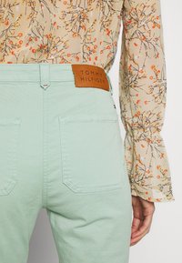 Miętowe spodnie z bawełnianej tkaniny z tylnymi kieszeniami; na nich znajduje się brązowa skórzana metka z napisem "TOMMY HILFIGER". Na górze widoczna kwiatowa bluzka.