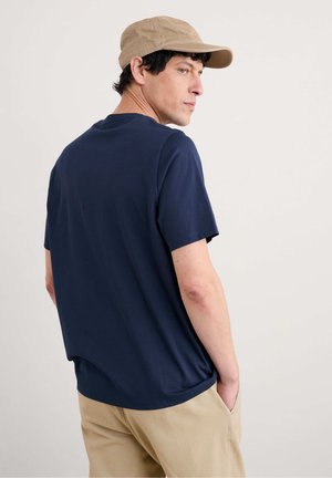 Hombre con gorra beige, camiseta azul marino y pantalones beiges de pie con las manos en los bolsillos, mirando por el hombro contra un fondo liso.